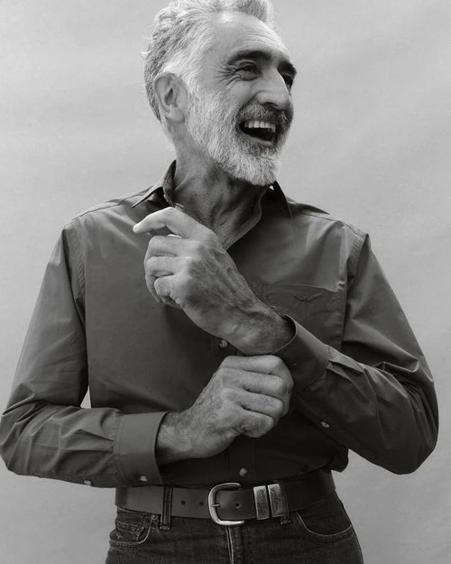 Elderly man with grey hair and beard laughing while adjusting his button-down shirt cuff.