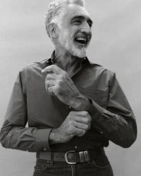 Elderly man with grey hair and beard laughing while adjusting his button-down shirt cuff.