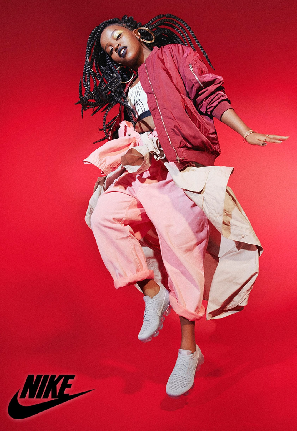 A dynamic fashion shot showcases a Black woman in a red jacket and pink pants, mid-air with Nike shoes, against a bold red backdrop.