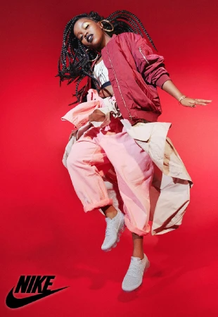 A dynamic fashion shot showcases a Black woman in a red jacket and pink pants, mid-air with Nike shoes, against a bold red backdrop.