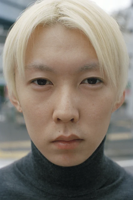Close-up portrait of a young adult with platinum blonde hair and a gray turtleneck sweater, set against a blurred outdoor background.