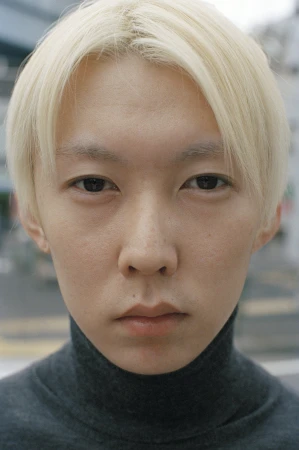 Close-up portrait of a young adult with platinum blonde hair and a gray turtleneck sweater, set against a blurred outdoor background.