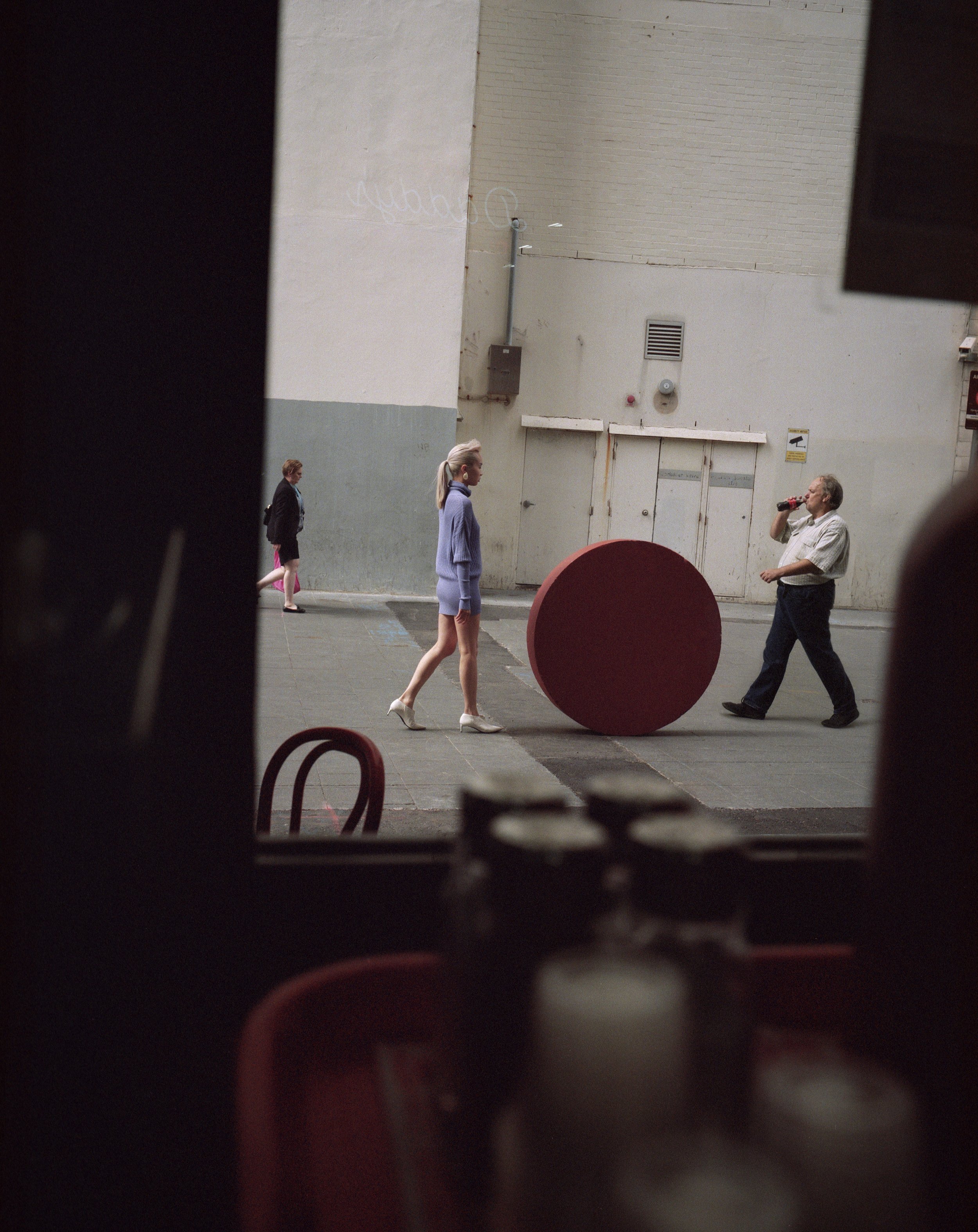 Pedestrians walk on a city street alongside a large red circle, as seen from an interior space with obscured objects in the foreground.