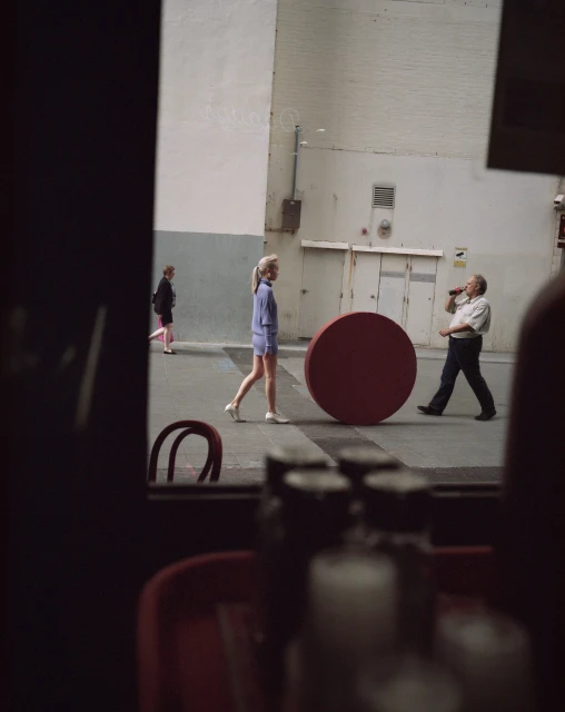 Pedestrians walk on a city street alongside a large red circle, as seen from an interior space with obscured objects in the foreground.