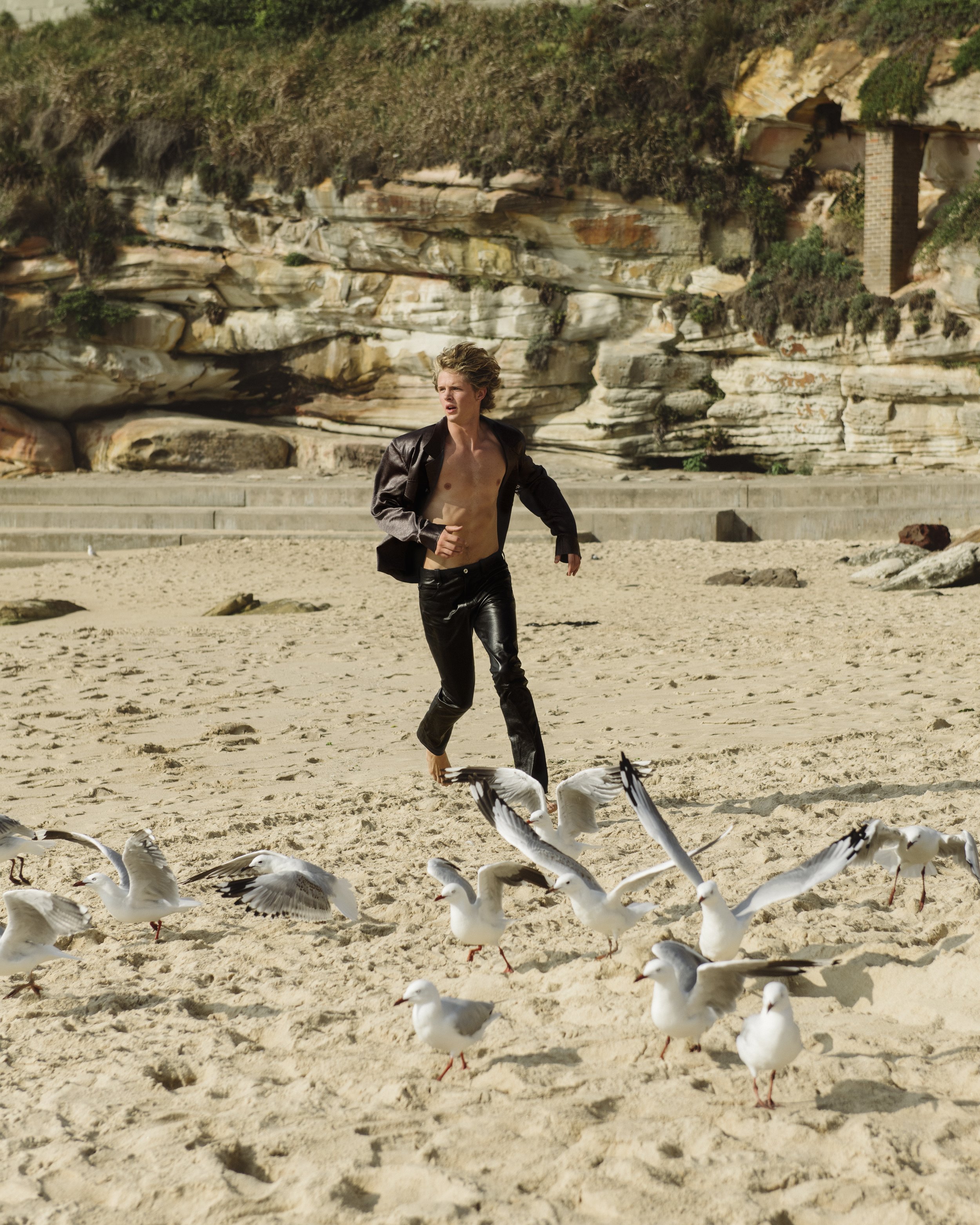 A young blond man runs barefoot on a beach as seagulls take flight. He wears leather pants and jacket, the background featuring a rocky cliffside.