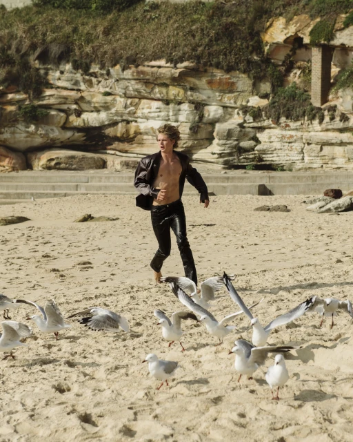 A young blond man runs barefoot on a beach as seagulls take flight. He wears leather pants and jacket, the background featuring a rocky cliffside.