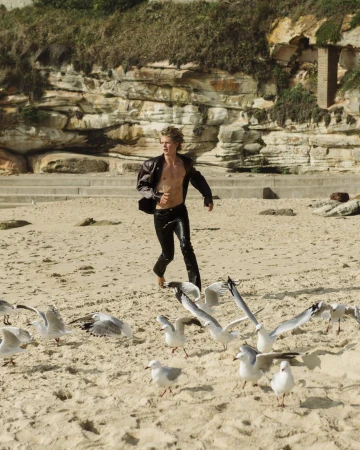 A young blond man runs barefoot on a beach as seagulls take flight. He wears leather pants and jacket, the background featuring a rocky cliffside.