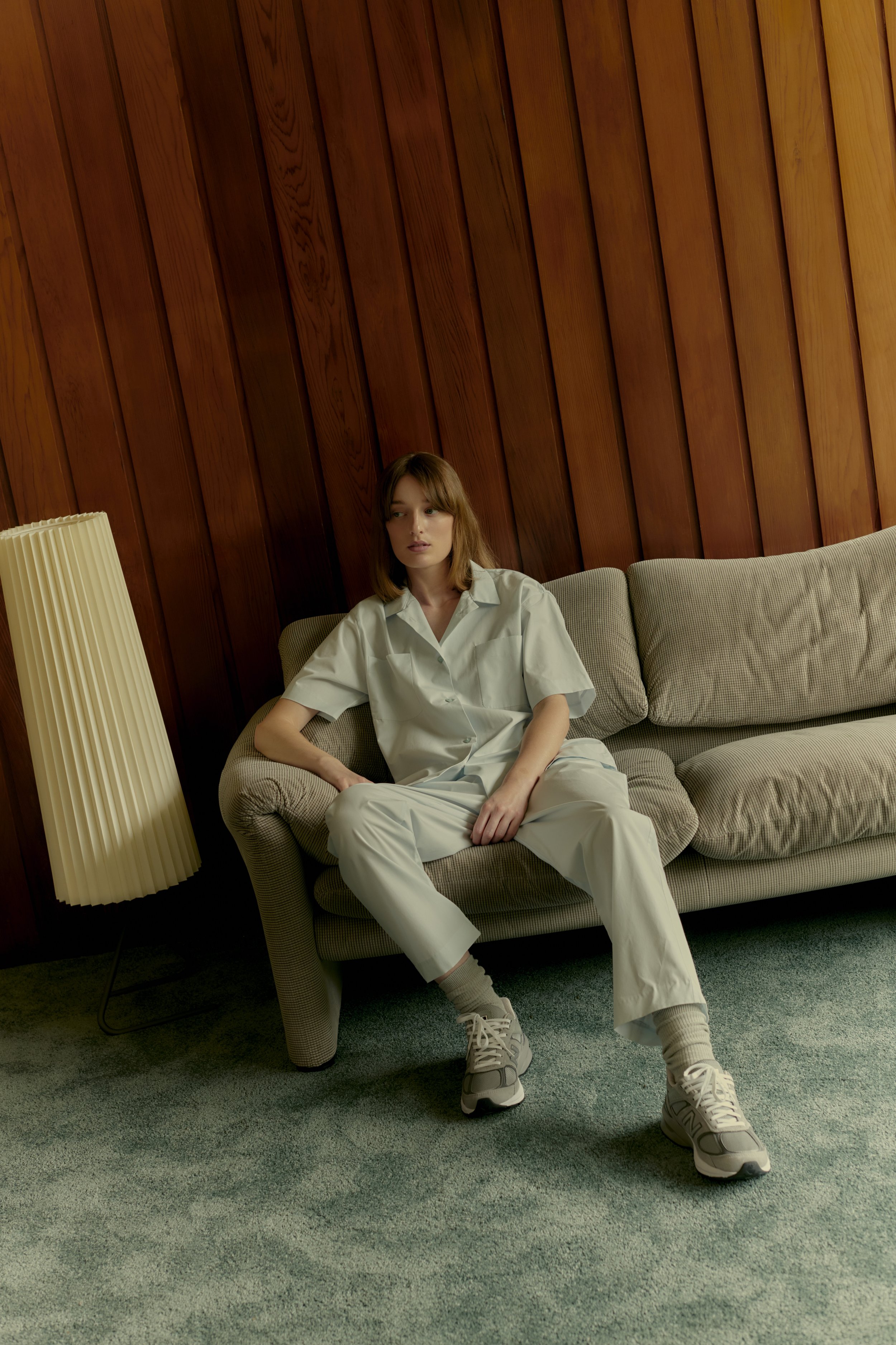 Woman in light blue collared shirt and pants sits on a gray couch in a room with wood paneled walls and a blue shag carpet.