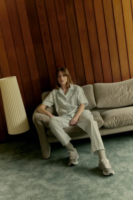 Woman in light blue collared shirt and pants sits on a gray couch in a room with wood paneled walls and a blue shag carpet.