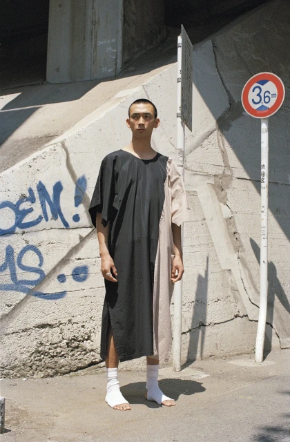 An Asian male poses in a color-blocked dress and white socks against a graffiti-covered concrete background with a road sign and an overpass.