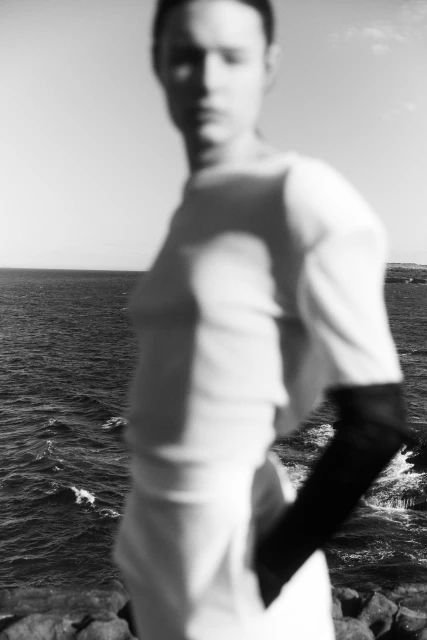 Monochrome fashion shot of an unfocused adult woman in a white dress standing before the ocean on a sunny day with rocky terrain.
