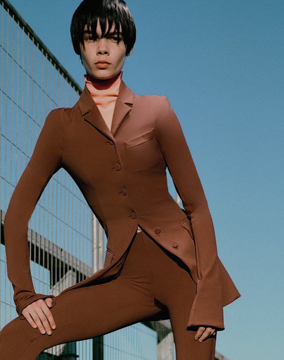Woman in brown suit poses against a blue sky and metallic structure. Fashion shot, full figure, natural light.