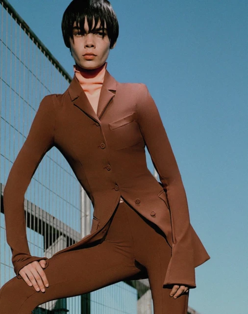 Woman in brown suit poses against a blue sky and metallic structure. Fashion shot, full figure, natural light.