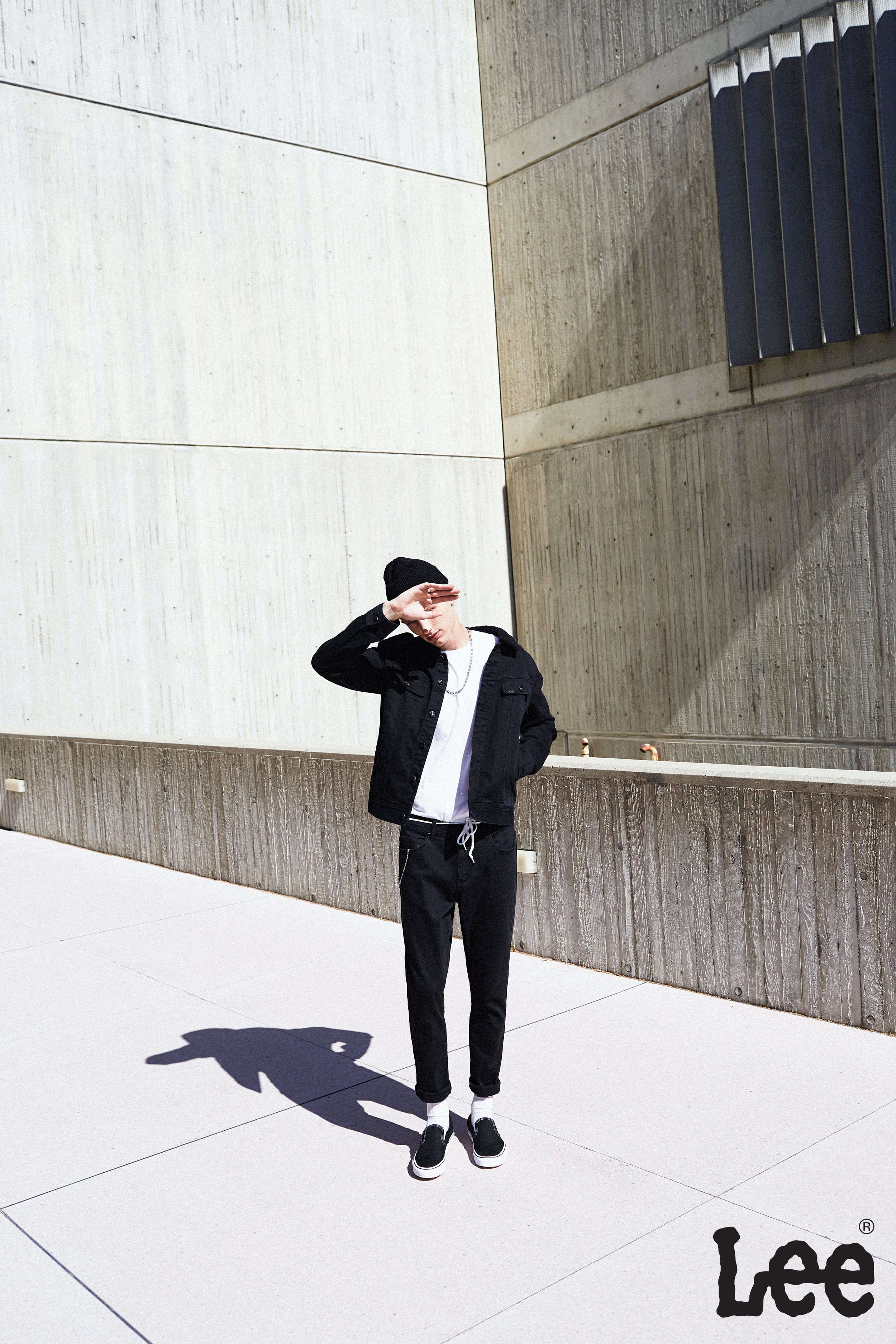 Man in black jacket and beanie poses against concrete walls, casting shadow on a sunny day, branded with Lee logo at bottom right.
