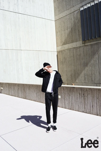 Man in black jacket and beanie poses against concrete walls, casting shadow on a sunny day, branded with Lee logo at bottom right.