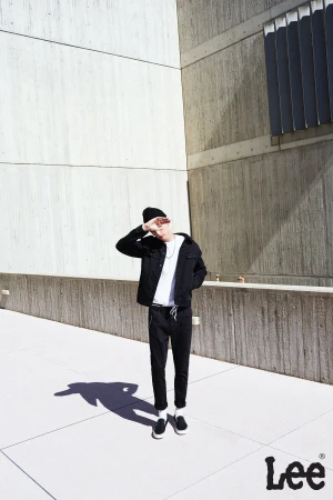 Man in black jacket and beanie poses against concrete walls, casting shadow on a sunny day, branded with Lee logo at bottom right.
