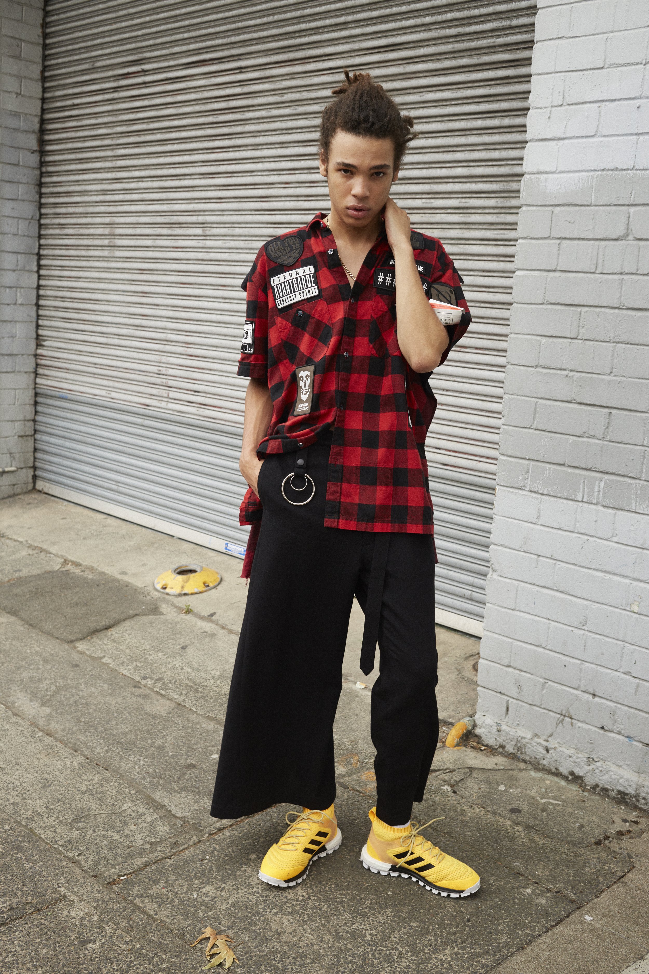 Young man stands on a city sidewalk wearing a red and black plaid shirt, black pants and yellow Adidas sneakers in an outdoor shot.