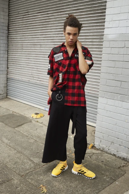 Young man stands on a city sidewalk wearing a red and black plaid shirt, black pants and yellow Adidas sneakers in an outdoor shot.