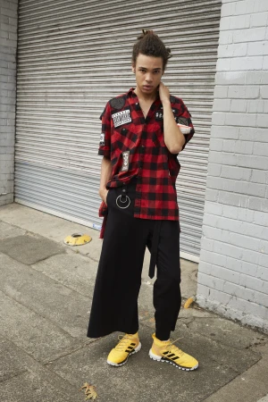 Young man stands on a city sidewalk wearing a red and black plaid shirt, black pants and yellow Adidas sneakers in an outdoor shot.