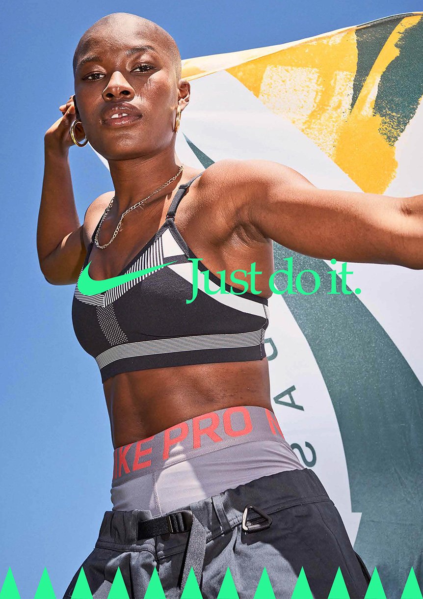 Athletic woman poses outdoors in Nike sports bra and athletic pants, holding a patterned flag, with blue sky in the background.