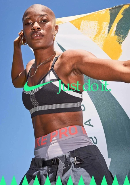 Athletic woman poses outdoors in Nike sports bra and athletic pants, holding a patterned flag, with blue sky in the background.