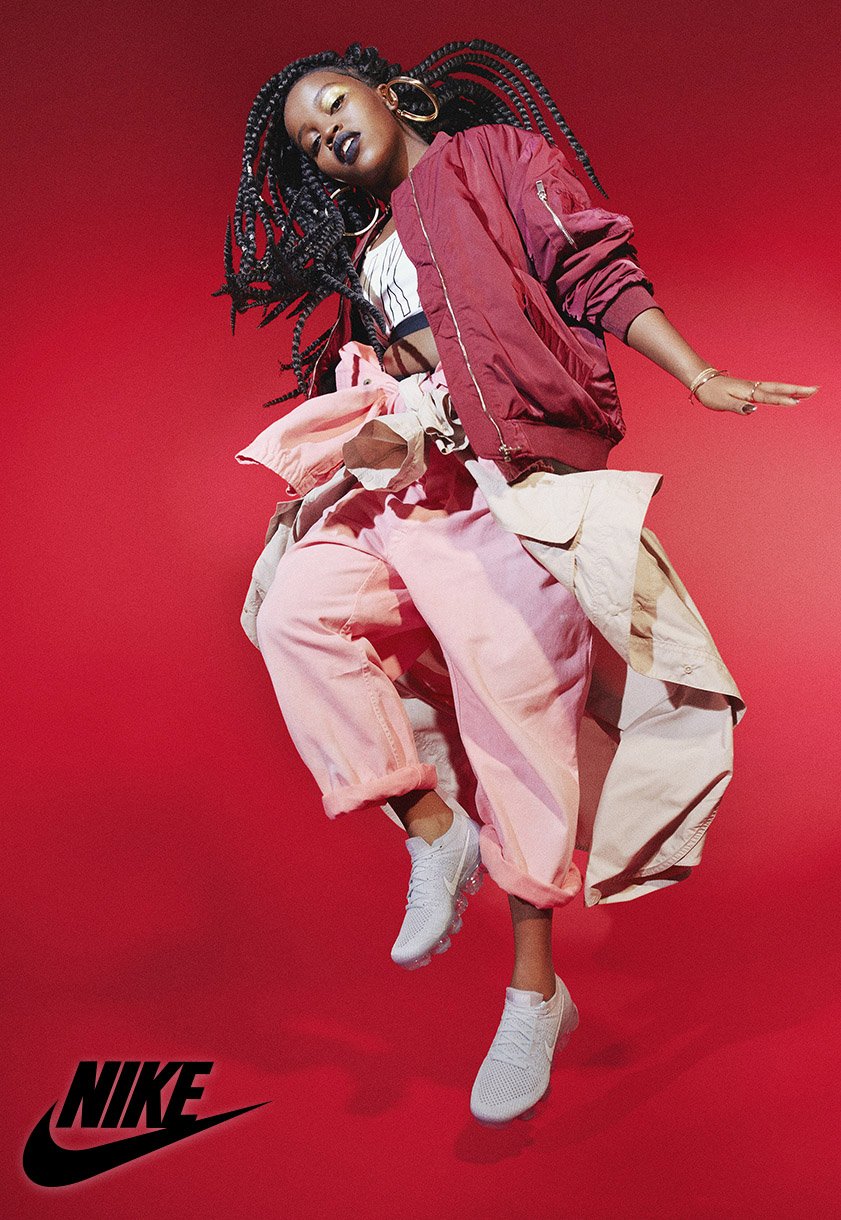 Full-length shot of an African American woman in streetwear jumping against a red background, showcasing a Nike logo.
