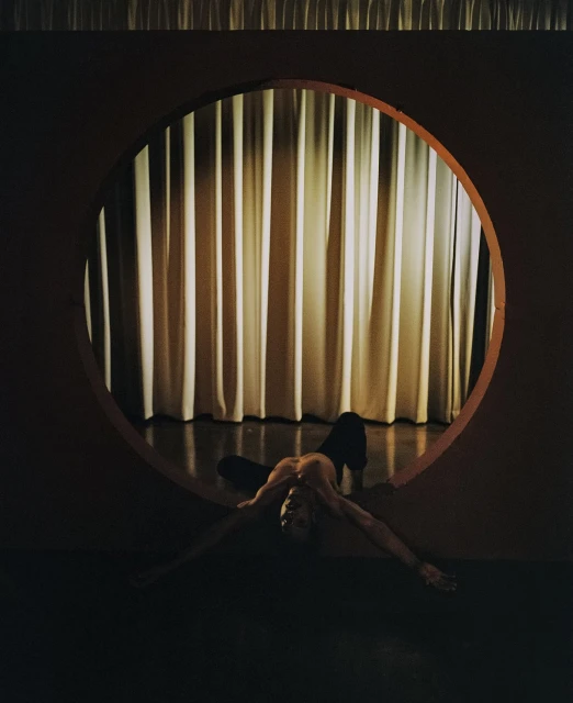 An adult is lying on the floor framed by a circle cut into a wall, with cream-colored drapes visible through the opening.