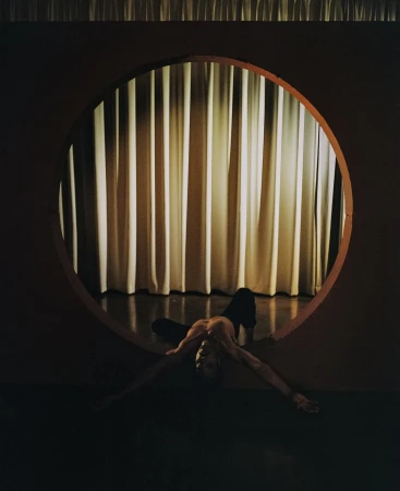 An adult is lying on the floor framed by a circle cut into a wall, with cream-colored drapes visible through the opening.