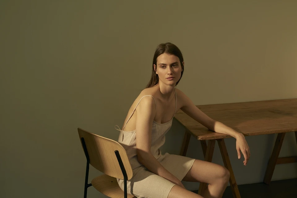 A woman in a light-colored dress sitting on a chair next to a wooden table, gazing away from the camera in a relaxed pose.
