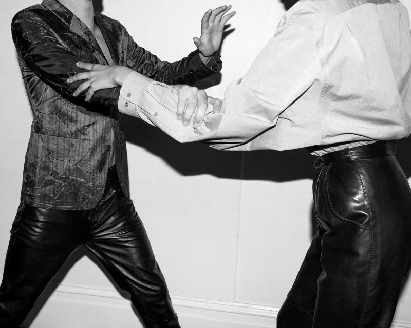 Monochrome fashion shot of two adults in leather pants and blazers, posed against a white wall with dramatic lighting and shadows.