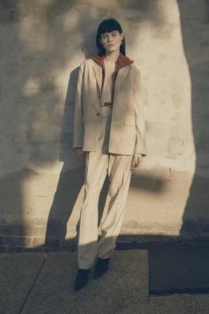 Model standing outdoors wearing a beige suit and rust top against a sunlit textured brick wall
