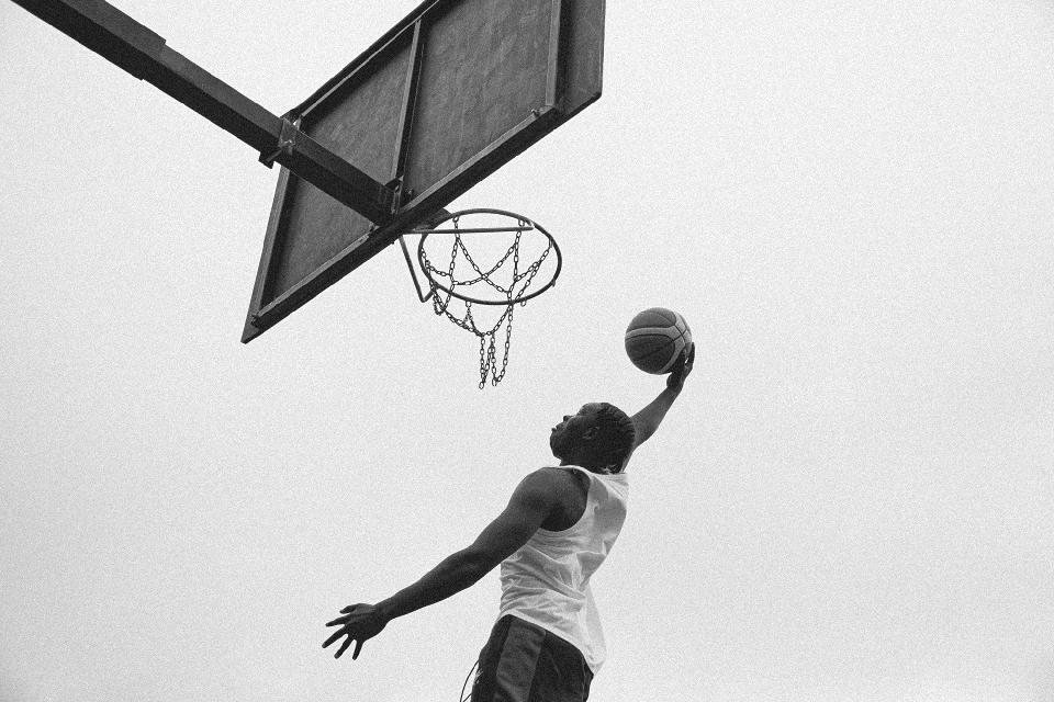 Monochrome image of an adult male basketball player in mid-air about to dunk a basketball in an outdoor setting with a cloudy sky.