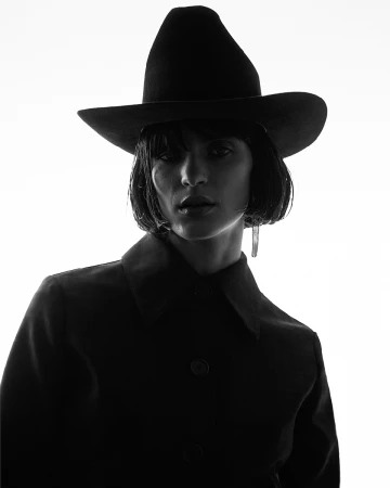 Monochrome portrait of a person wearing a wide-brimmed hat and dark collared jacket against a bright white background.