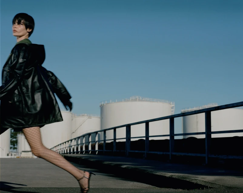 A stylish woman in a black leather coat is walking past large white storage tanks with blue sky above, creating a fashion editorial image.