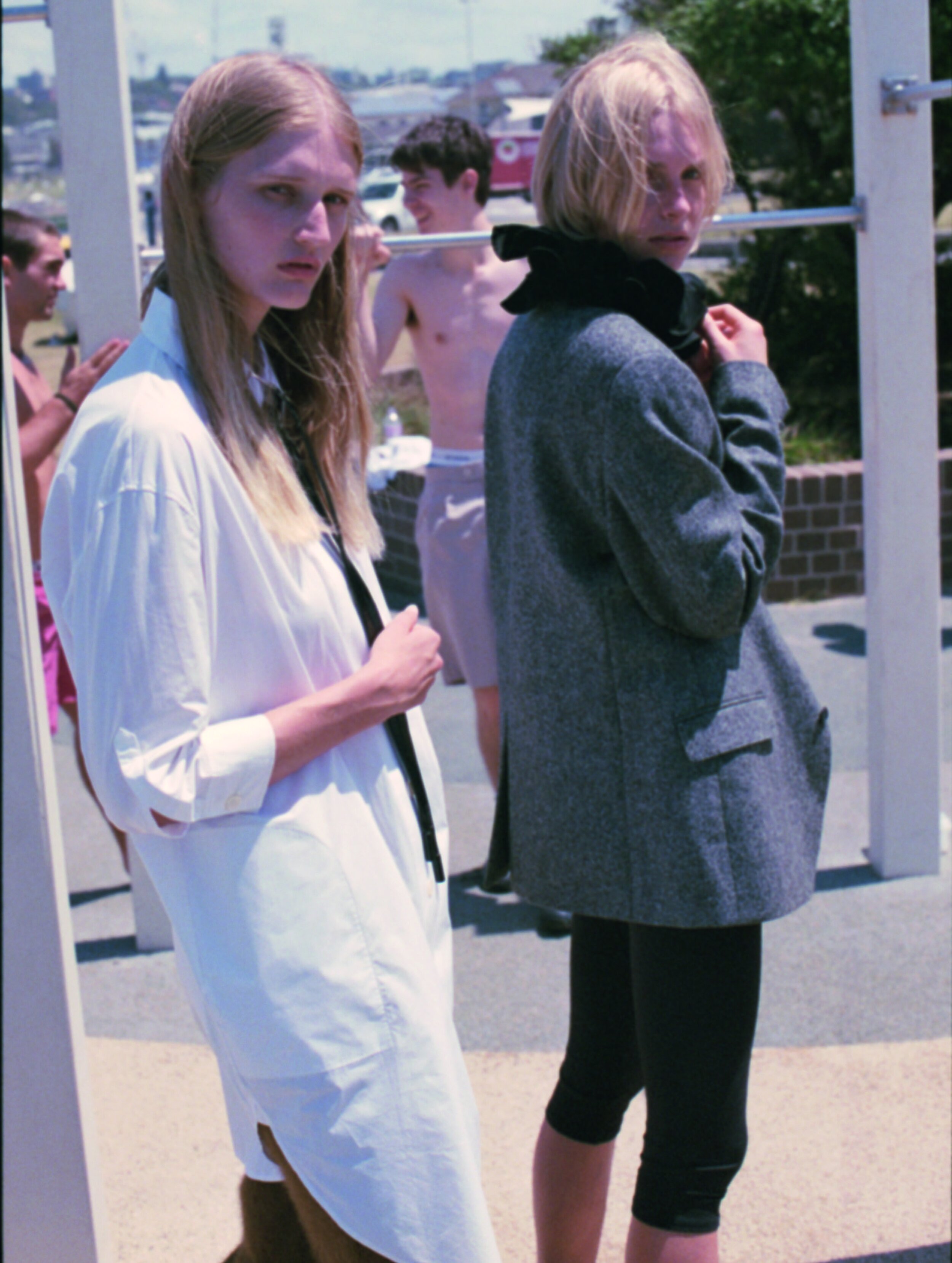 Two fair-skinned adults in a casual fashion shot, outdoors during the day. One in white shirt dress, the other in a grey jacket with leggings.