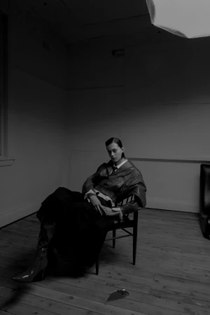 A dark-haired female model sitting on a wooden chair in a room with a wooden floor and white walls, styled by Nicki Colbran.