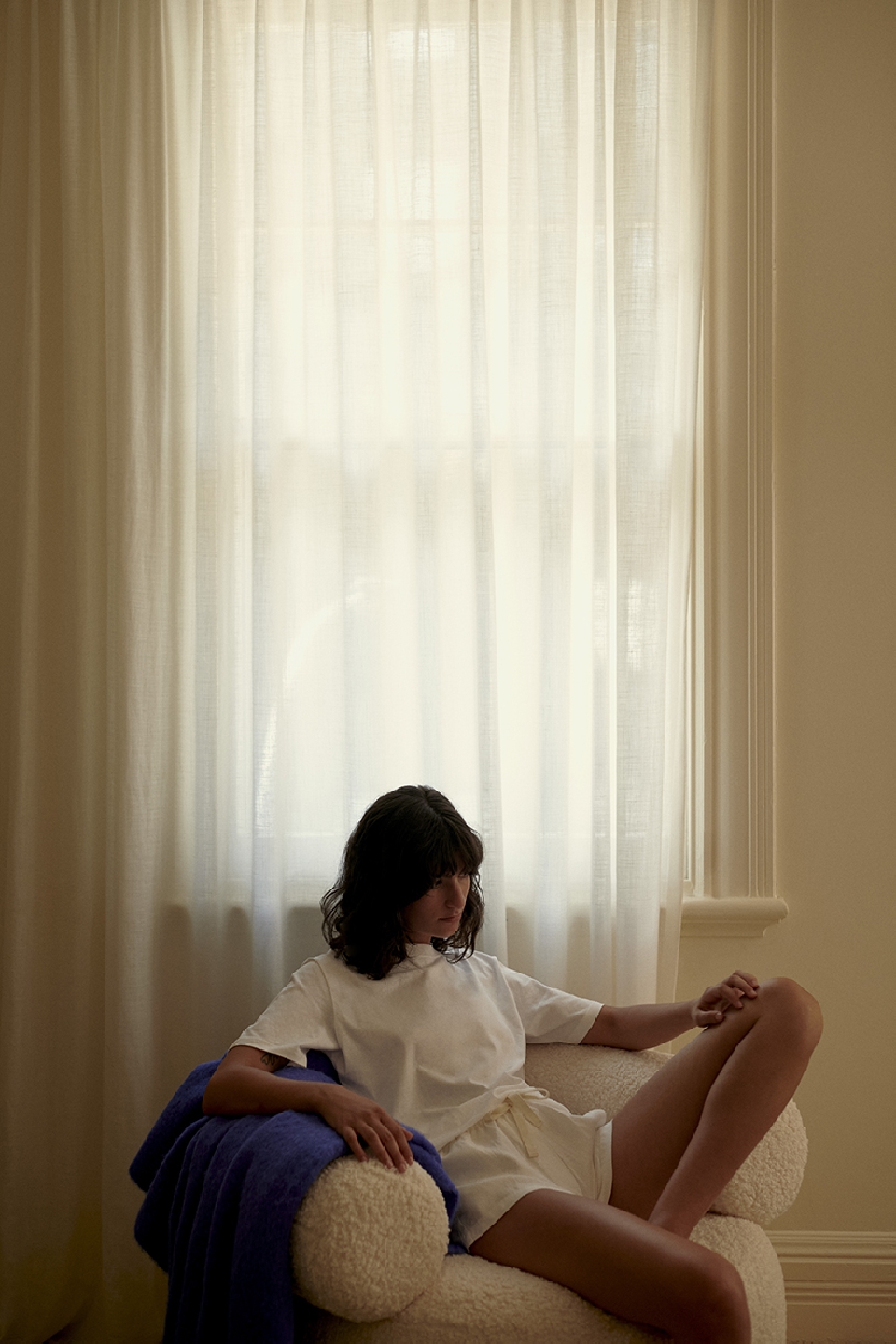 Brunette woman poses in a white t-shirt and shorts, seated on a textured white chair indoors with a blue blanket and window.