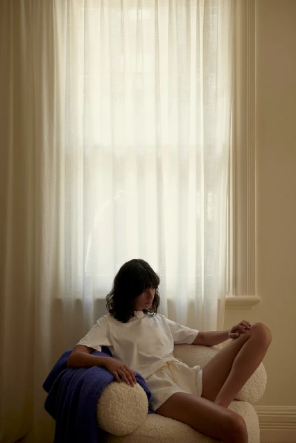 Brunette woman poses in a white t-shirt and shorts, seated on a textured white chair indoors with a blue blanket and window.