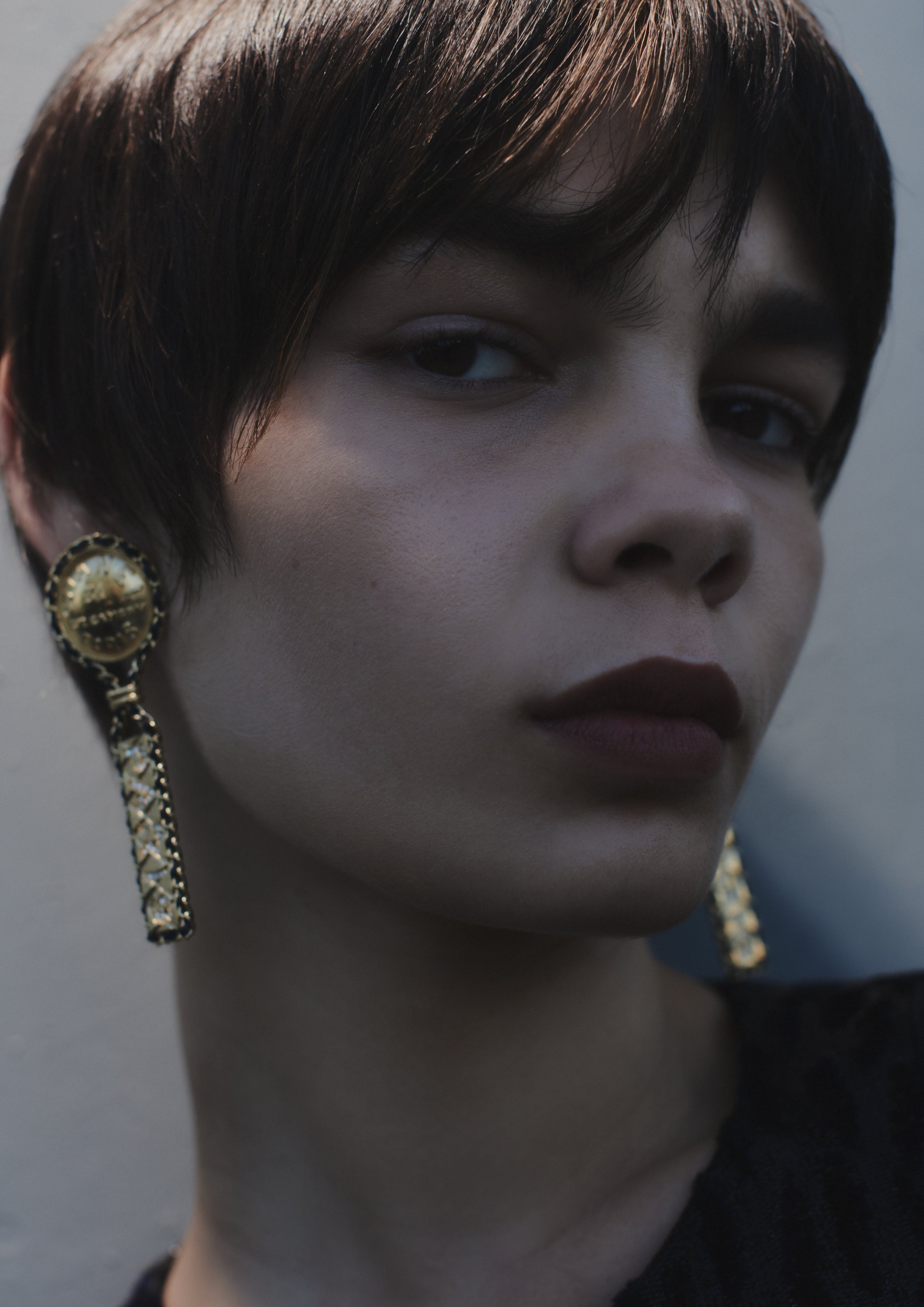 Close-up portrait of a young woman with dark brown short hair, wearing large gold statement earrings, against a neutral background.