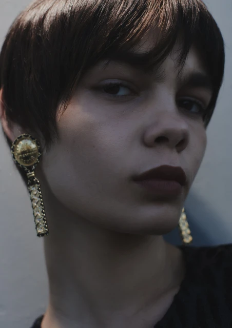 Close-up portrait of a young woman with dark brown short hair, wearing large gold statement earrings, against a neutral background.