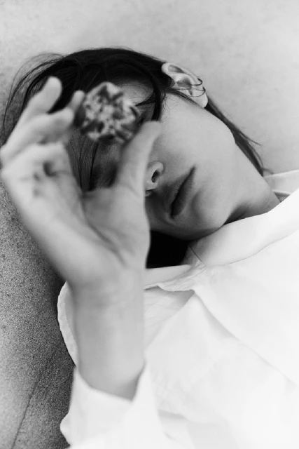 Black and white portrait of a woman lying down, holding a textured object in front of her face, wearing a collared shirt.