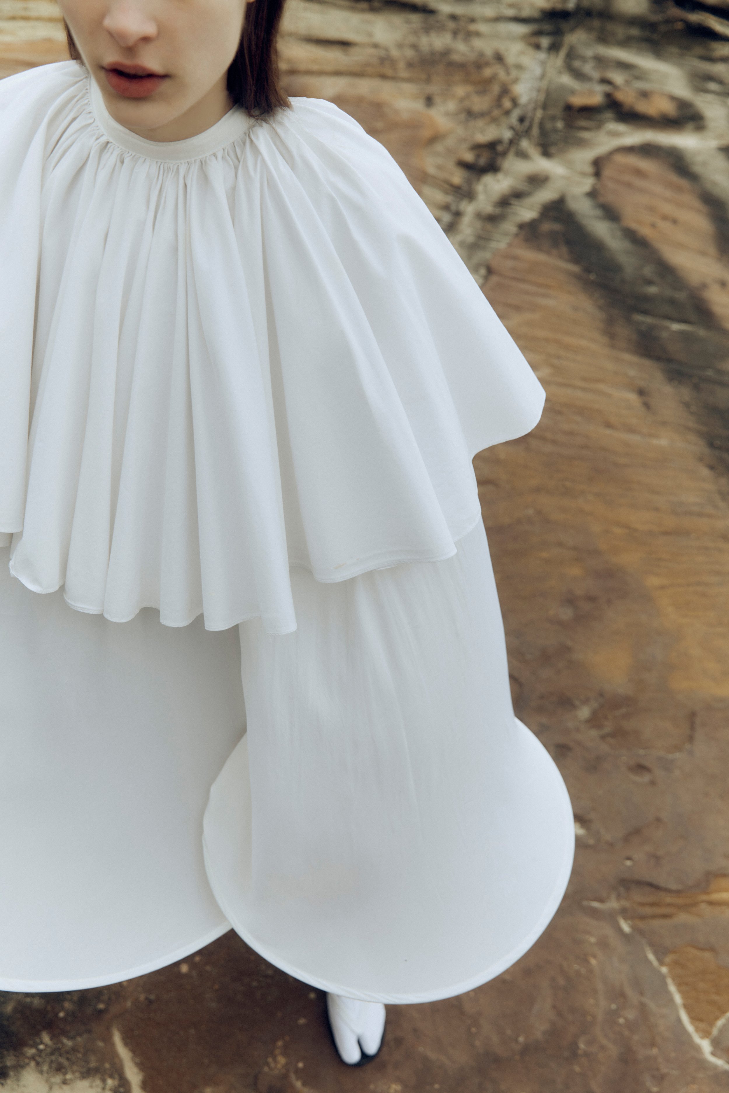 Overhead shot of a slender person in a flowing, white, two-tiered dress standing on a textured, tan-and-brown surface.