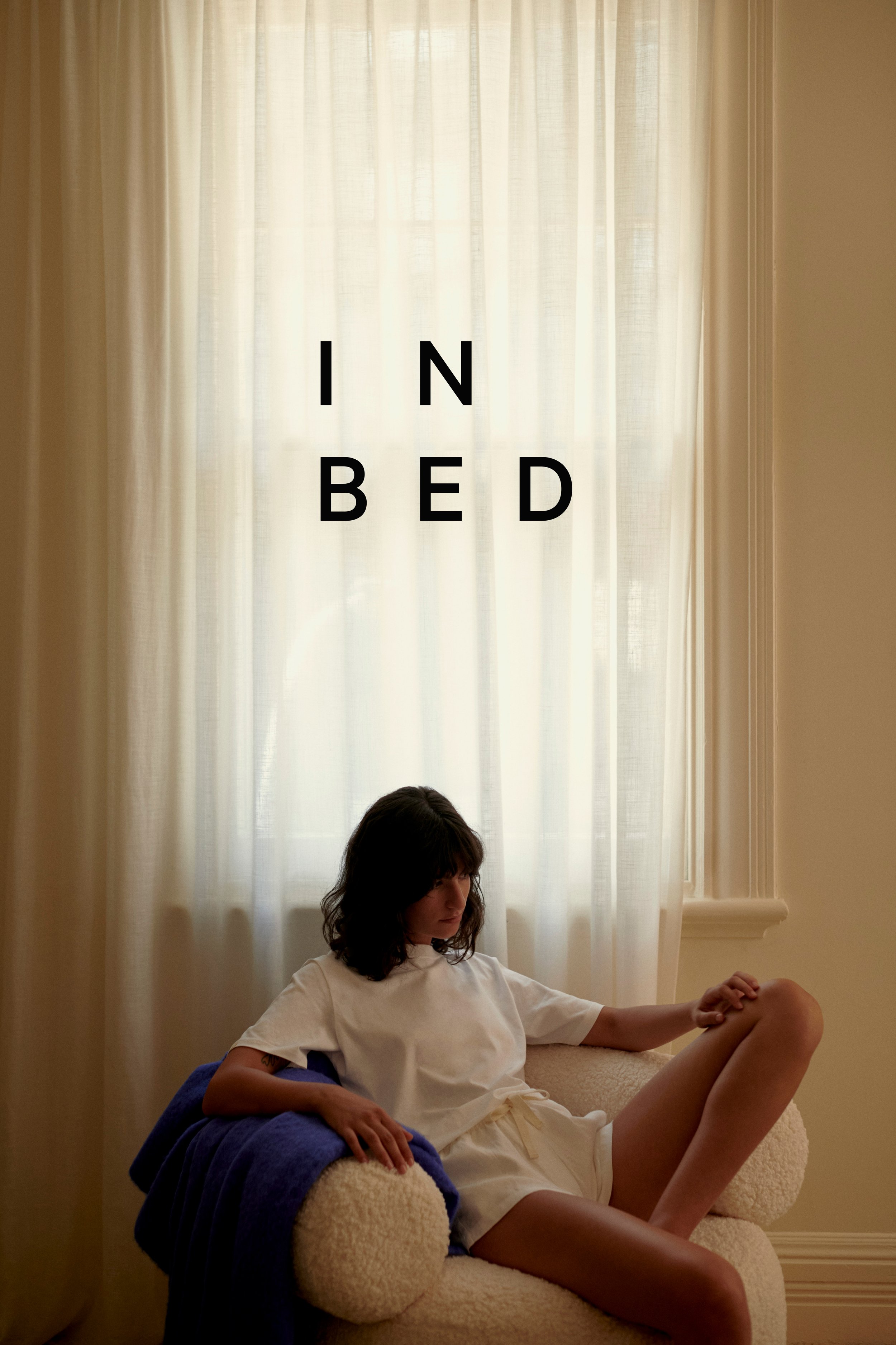Woman in white shirt and shorts sits in a chair near a window with white curtains in an indoor setting with soft, neutral lighting.