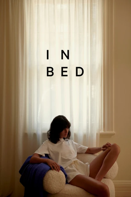 Woman in white shirt and shorts sits in a chair near a window with white curtains in an indoor setting with soft, neutral lighting.