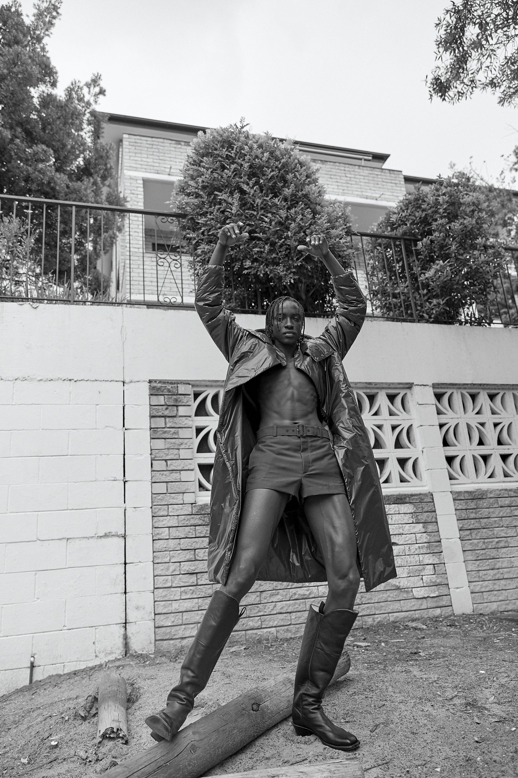 Black and white portrait of a male model wearing a belted coat and tall boots, styled by Nicki Colbran in Sydney.
