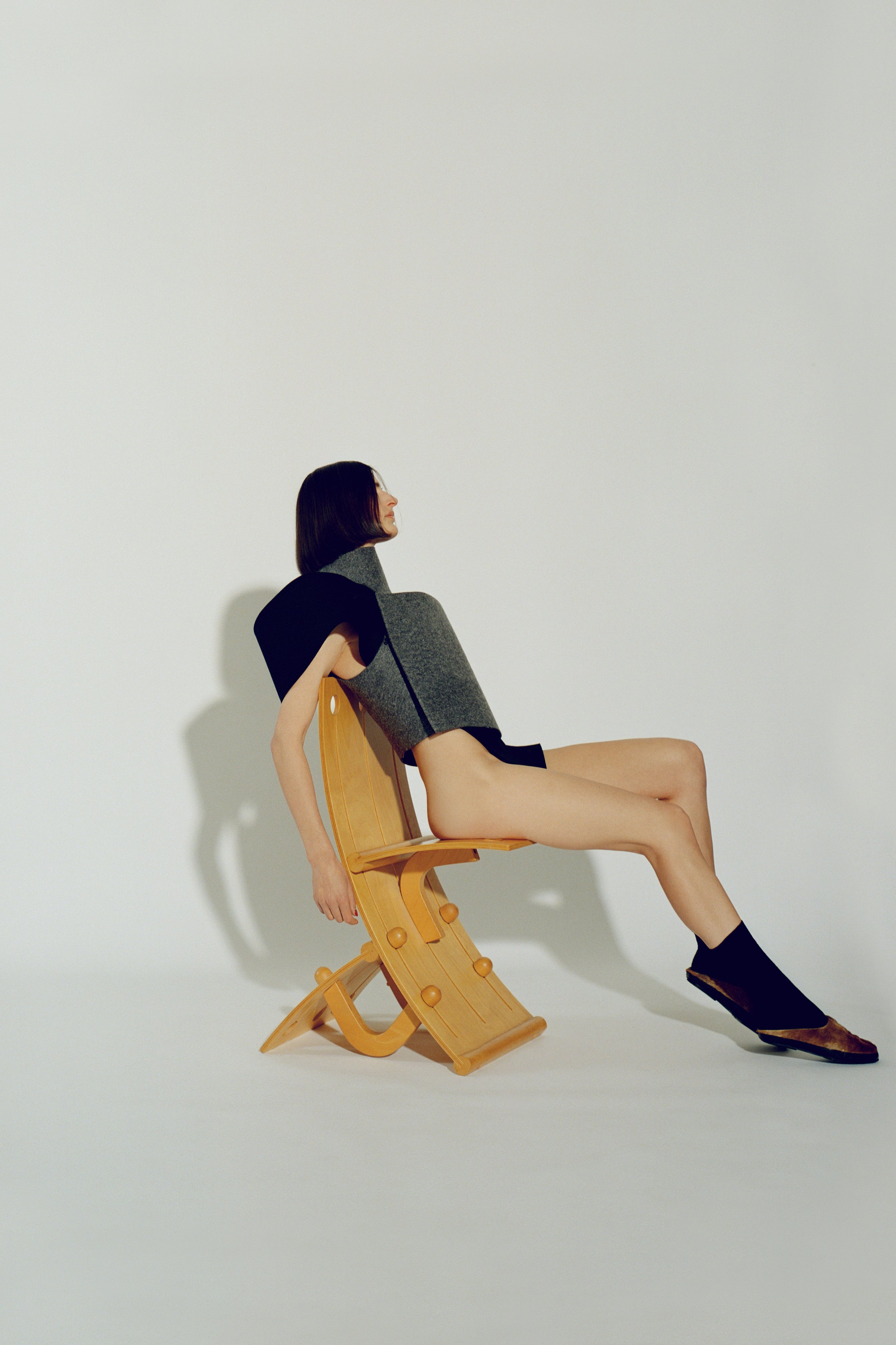 Fashion shot of a woman with short dark hair wearing a grey top and black sock-shoes, posed in a pale yellow chair against white background.