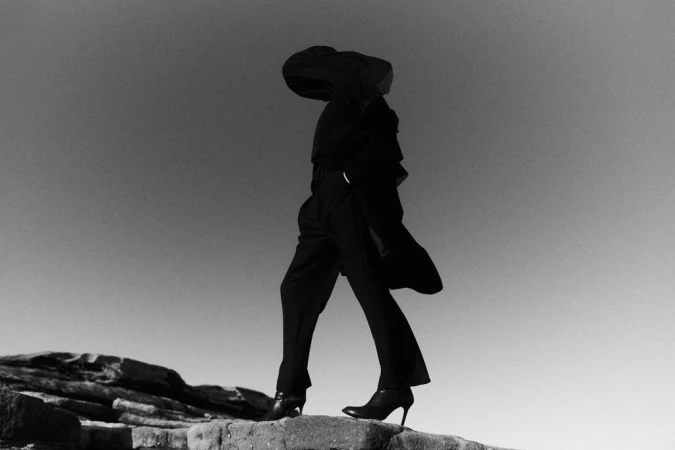 Monochrome fashion image of a model wearing dark trousers and heeled boots posing outdoors on rocky terrain under an open sky.
