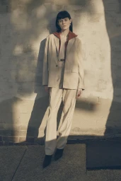 Model standing outdoors wearing a beige suit and rust top against a sunlit textured brick wall