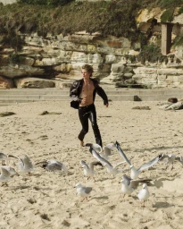 A young man running barefoot on a sandy beach amidst a flock of scattered seagulls with rocky cliffs behind.