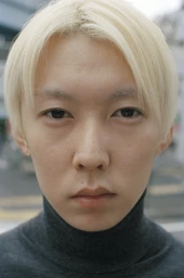 Close-up portrait of a person with bleached blonde parted hair and dark turtleneck shirt against an out-of-focus street background
